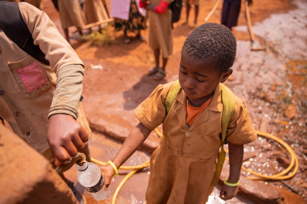Little african schoolgirl collects water in the bottle from the tap of the fountain